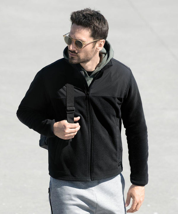 A man wearing sunglasses, a black jacket, grey sweatpants, and carrying a Nimbus NP17M over his shoulder walks on a concrete surface.