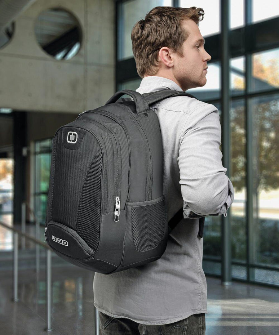 A man in a light gray shirt is standing indoors carrying an Ogio OG001 with multiple zippered compartments and mesh pockets. The background shows large windows and a modern interior.