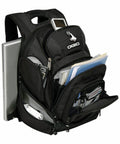A black Ogio OG002 backpack with multiple compartments is shown. It holds a laptop, papers, files, a smartphone, and various small items, indicating ample storage and organizational features.