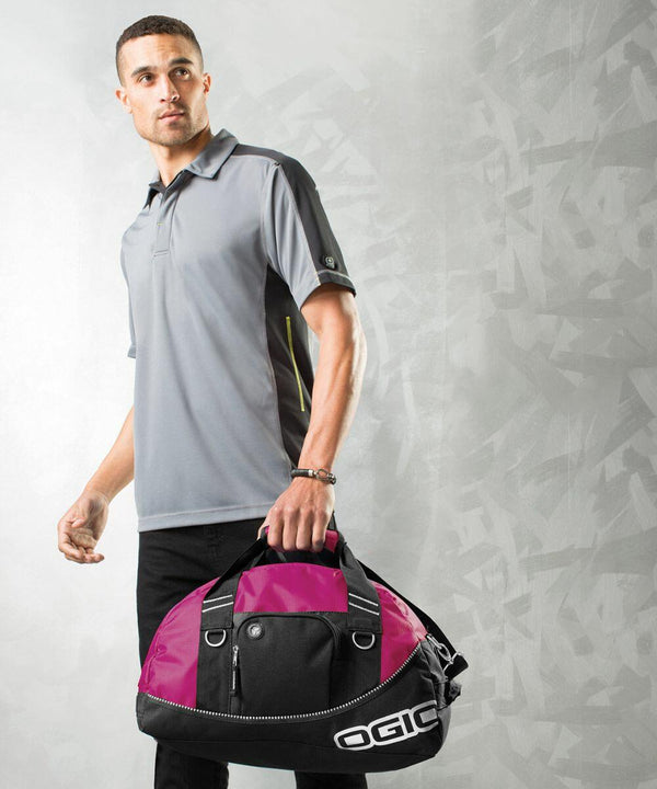 A man in a gray polo shirt and black pants holds a black and pink Ogio OG010 duffel bag against a gray textured background.