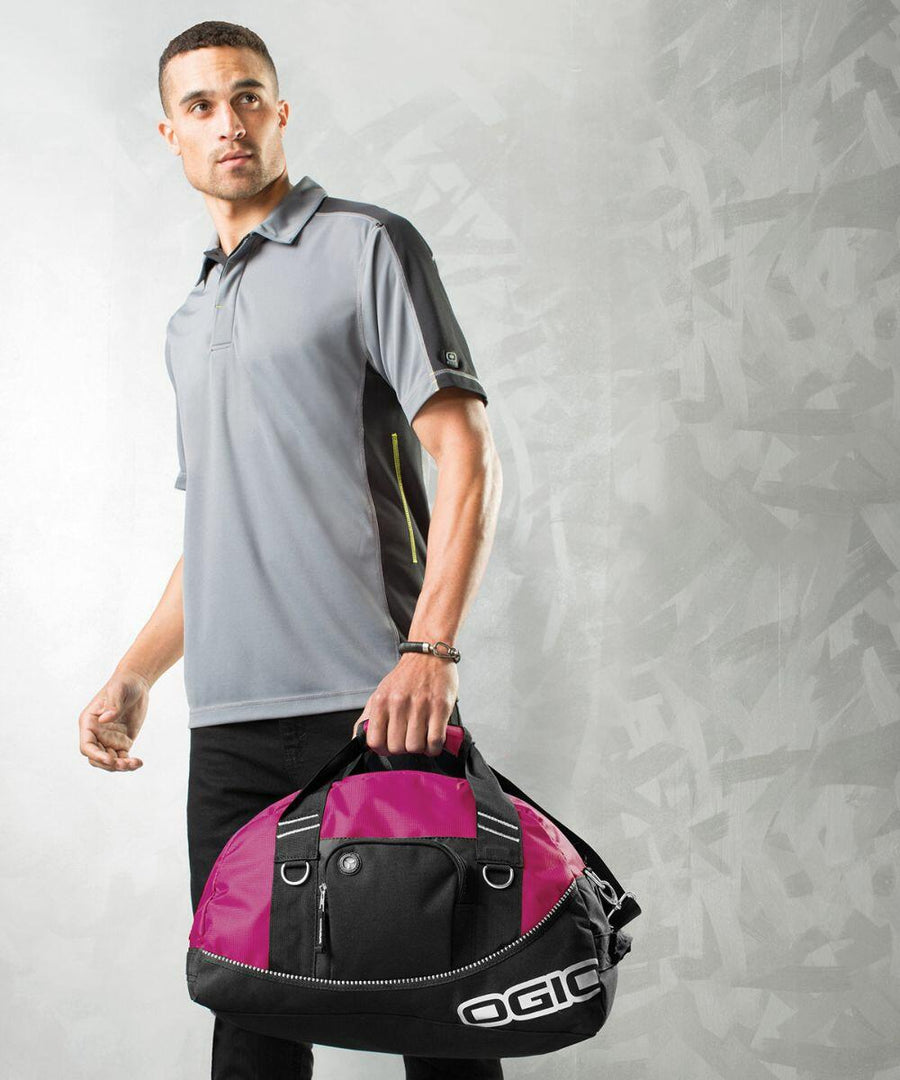 A man in a gray polo shirt and black pants holds a black and pink Ogio OG010 duffel bag against a gray textured background.