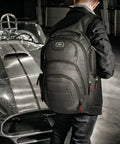 A person wearing a black jacket and an Ogio OG018 backpack stands next to a vintage sports car with a racing stripe.