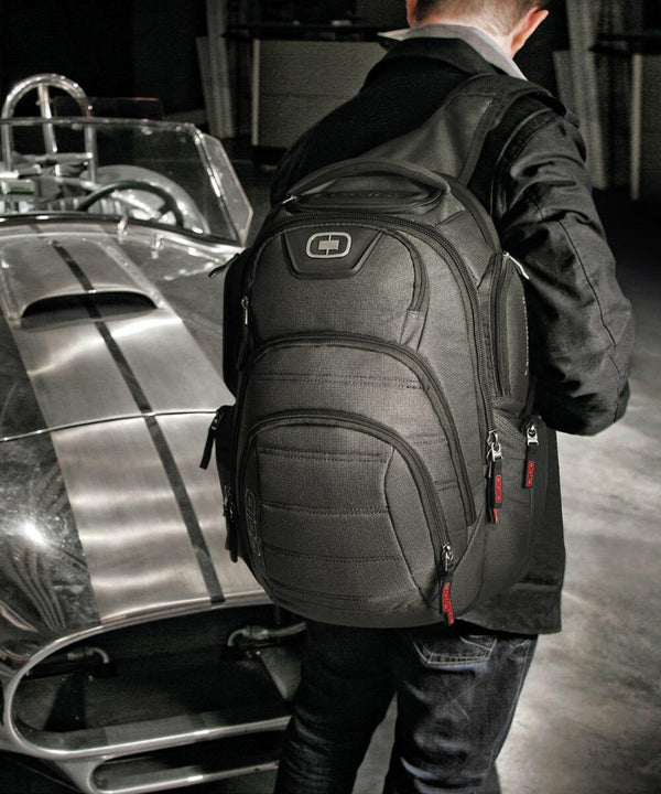 A person wearing a black jacket and an Ogio OG018 backpack stands next to a vintage sports car with a racing stripe.
