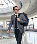 A man in a blazer, tie, and sunglasses walks confidently through a modern airport terminal carrying an Ogio OG027 backpack.