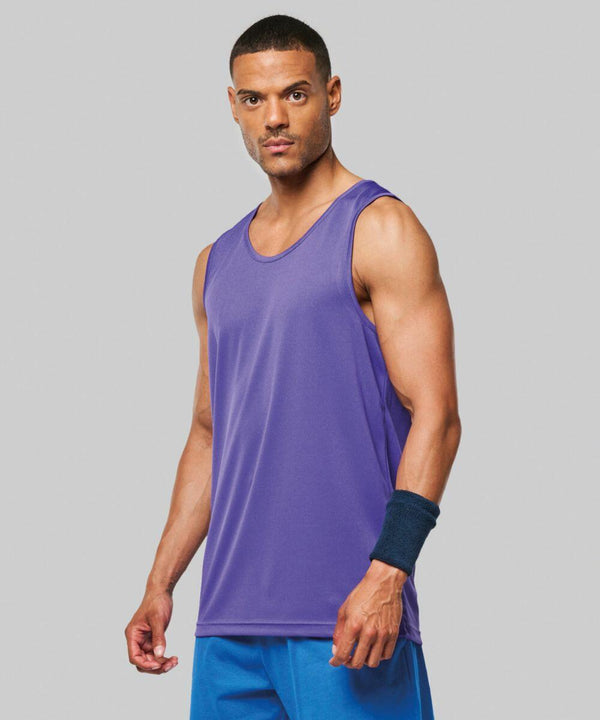 A man wearing a purple PA441 tank top by Kariban Proact, blue shorts, and a blue wristband stands against a plain gray background.