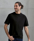 A man with long dark hair tied back stands against a light grey concrete wall, wearing the Premier Utility 2.0 oversized t-shirt in black and dark trousers, looking to the side and smiling.