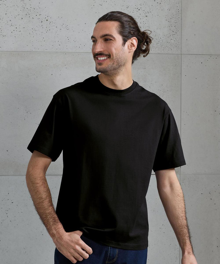 A man with long dark hair tied back stands against a light grey concrete wall, wearing the Premier Utility 2.0 oversized t-shirt in black and dark trousers, looking to the side and smiling.