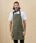 A man with long hair tied back wears a white t-shirt, blue patterned neckerchief, and the Premier Utility 2.0 ripstop apron in green. He stands before a light wood background, holding a small white object in his right hand.