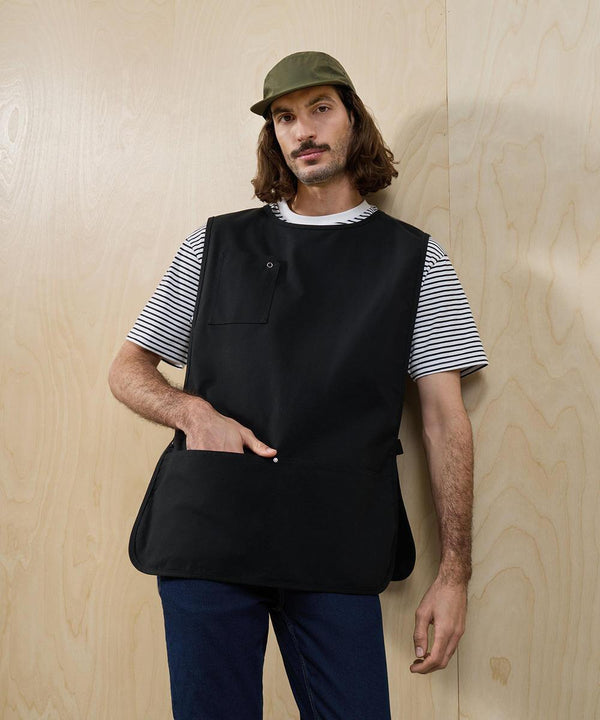 A person with long brown hair and a moustache wears Premier's Utility 2.0 vest apron over a striped shirt, green cap, and blue trousers, standing against a light wood-panelled wall with one hand in the apron pocket.