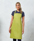 A woman stands in front of a neutral background, wearing a Premier PR102 lime green apron over a dark short-sleeve shirt.