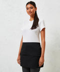 A woman stands against a textured white wall, wearing a white short-sleeve shirt and a black Premier PR105 apron. She has long dark hair and a neutral expression.