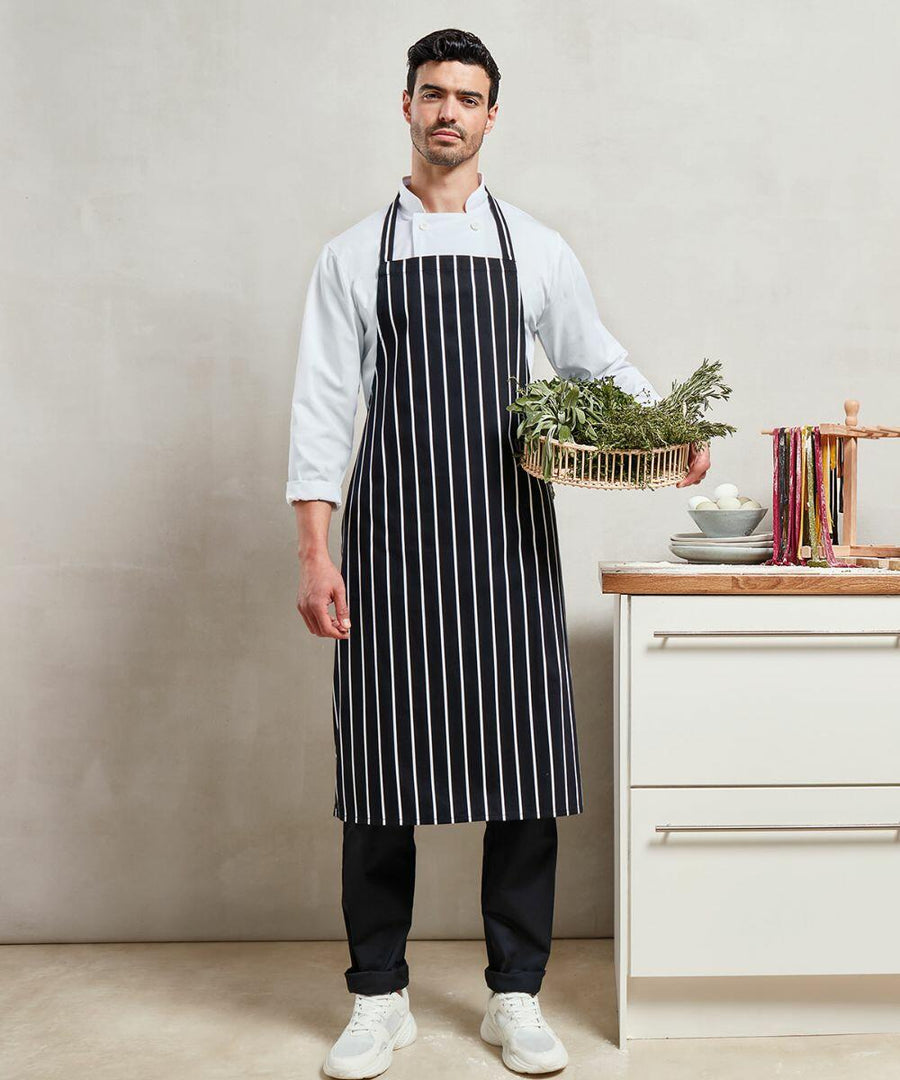 A man in a black and white striped apron stands in a kitchen, holding a basket of greens. He is next to a counter with Premier PR110 pasta and eggs.