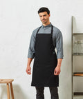 Man wearing a grey Premier PR112 button-up shirt and a black apron, standing in front of a white textured wall with shelves and stools in the background.