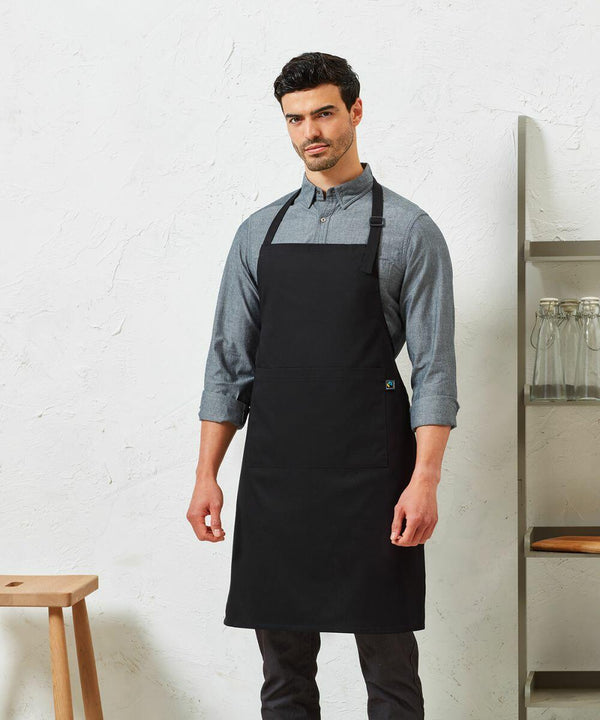 Man wearing a grey Premier PR112 button-up shirt and a black apron, standing in front of a white textured wall with shelves and stools in the background.