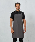 A man wearing a grey Premier PR115 apron over a black polo shirt stands against a light grey textured background. He is smiling and facing the camera.