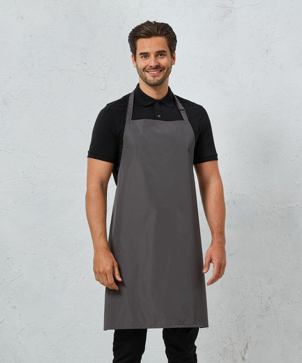 A man wearing a grey Premier PR115 apron over a black polo shirt stands against a light grey textured background. He is smiling and facing the camera.