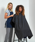 A hairstylist stands behind a client seated in a chair, wearing a Premier PR116 black cape, holding scissors, and preparing for a haircut in a salon.