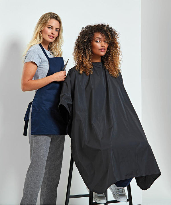 A hairstylist stands behind a client seated in a chair, wearing a Premier PR116 black cape, holding scissors, and preparing for a haircut in a salon.