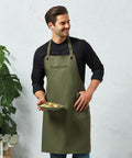 A person wearing a black Premier PR121 shirt and green apron stands holding a plate of food, with a plant in the background.