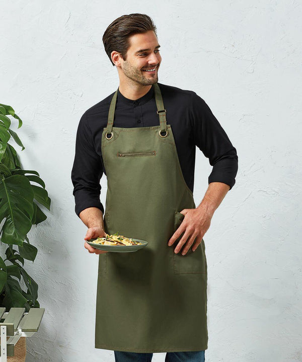 A person wearing a black Premier PR121 shirt and green apron stands holding a plate of food, with a plant in the background.