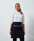 A woman stands against a textured white wall wearing a white collared shirt and a Premier PR128 denim apron.