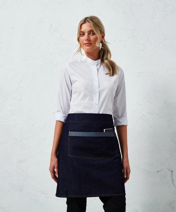 A woman stands against a textured white wall wearing a white collared shirt and a Premier PR128 denim apron.