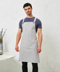 A man wearing a grey Premier apron, product PR129, over a blue t-shirt stands in front of a textured white wall next to a small table with a vase and plates.