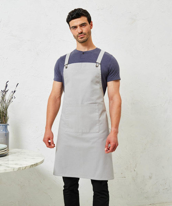 A man wearing a grey Premier apron, product PR129, over a blue t-shirt stands in front of a textured white wall next to a small table with a vase and plates.