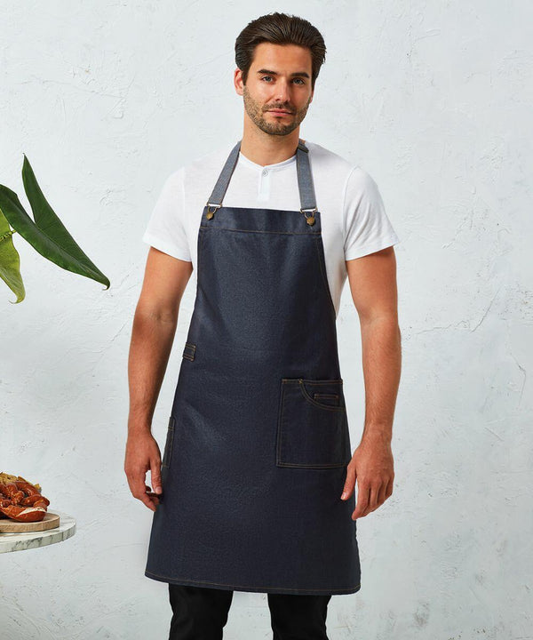 A man in a white T-shirt and Premier PR134 denim apron stands against a white textured wall, with a small table to the side holding a dish of food and a potted plant.