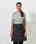 Person standing against a white textured wall, wearing a gray long-sleeve shirt and a black Premier PR135 apron with pockets, smiling and looking at the camera.