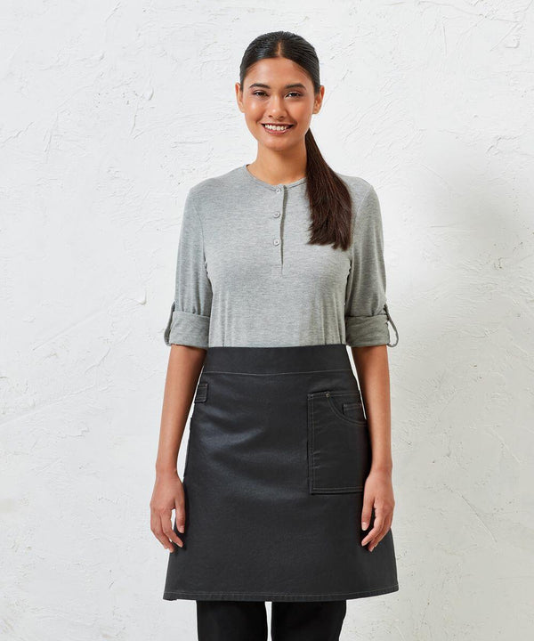 Person standing against a white textured wall, wearing a gray long-sleeve shirt and a black Premier PR135 apron with pockets, smiling and looking at the camera.