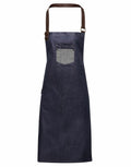 Introducing the Premier PR136, a dark blue denim apron featuring a leather neck strap and a small chest pocket. The pocket, enhanced with a lighter blue accent and secured by metal rivets, adds both style and functionality to this design.