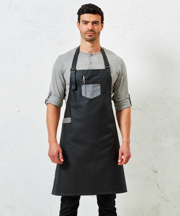 A man stands against a plain white background wearing a gray shirt and a black apron with a front pocket, which contains a pen. The apron is the PR136 by Premier.