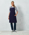 A person in a striped shirt, dark jeans, sneakers, and a navy Premier PR137 stands against a light-colored, textured wall with one hand in the apron pocket.