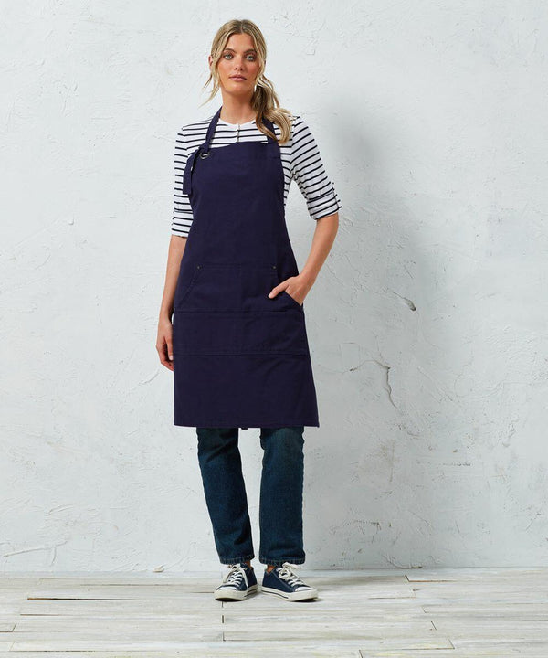 A person in a striped shirt, dark jeans, sneakers, and a navy Premier PR137 stands against a light-colored, textured wall with one hand in the apron pocket.