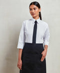 A woman in a white shirt, black tie, and black Premier PR141 apron with her left hand in the apron pocket, standing against a plain background.