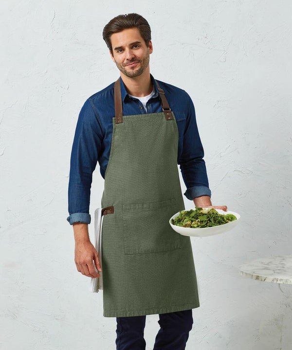 A man wearing a Premier PR144 and green apron holds a plate of salad. He stands against a light-colored wall and a marble table is partially visible.