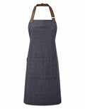 Introducing the Premier PR144: a sturdy denim apron featuring a front pocket and leather neck strap. Its simple design includes dark blue fabric complemented by two brown leather details at the neck, making it ideal for both cooking and crafting activities.