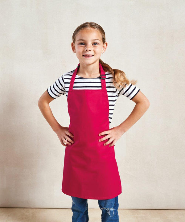 A young girl stands with hands on hips, wearing a striped shirt, blue jeans, and a bright red Premier PR149 apron, smiling at the camera.