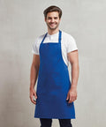 A man with short hair, wearing a white shirt and a blue apron labeled "Premier PR154," stands against a plain light background, smiling.
