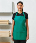 A person with dark hair pulled back, wearing a green apron over a black shirt, stands in front of a light-colored wall and a shelving unit with wooden boxes featuring the PR159 by Premier.