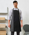 A man wearing a gray shirt and a Premier PR162 black apron stands in front of a white wall with shelves containing wooden trays and a gray chair nearby.