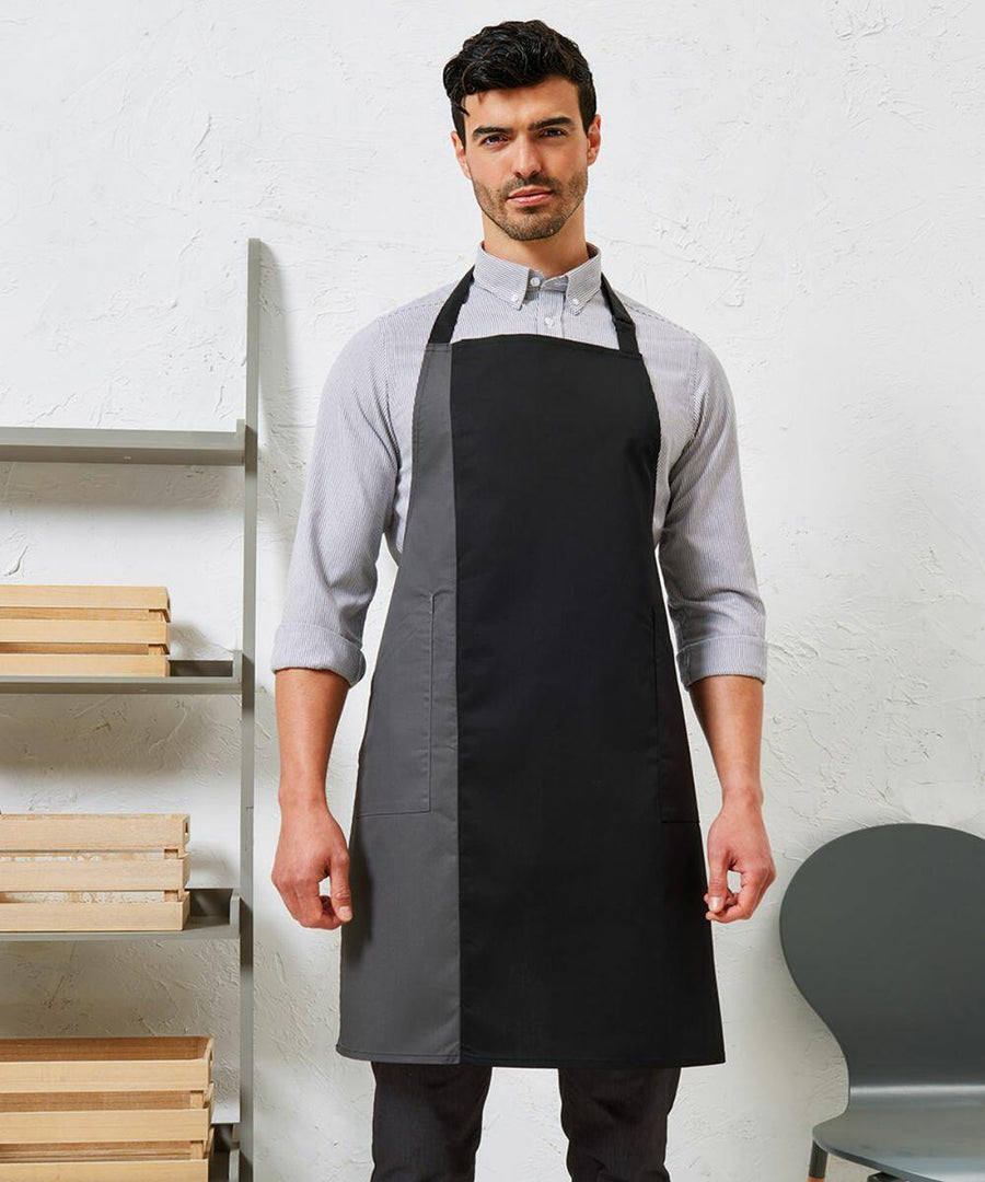 A man wearing a gray shirt and a Premier PR162 black apron stands in front of a white wall with shelves containing wooden trays and a gray chair nearby.