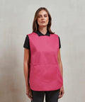 A woman is standing against a neutral background, wearing a black shirt and black pants with a bright pink sleeveless Premier PR171 apron. The apron has a large front pocket.