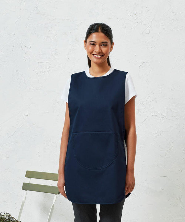 A person wearing a white shirt and a navy blue Premier PR172 with a front pocket stands in front of a plain white wall, smiling. A green chair is partially visible in the background.