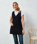 A person wearing a black Premier PR177 sleeveless apron over a white t-shirt is standing next to a wooden chair against a white textured wall.