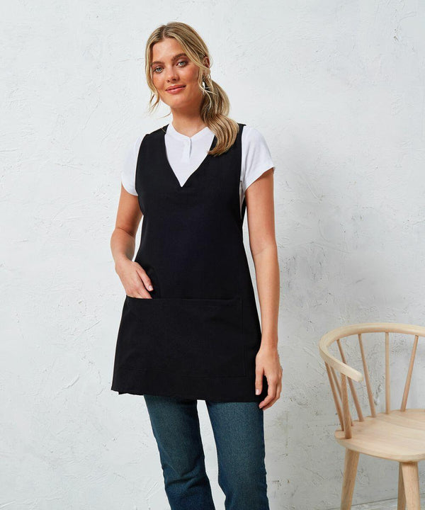 A person wearing a black Premier PR177 sleeveless apron over a white t-shirt is standing next to a wooden chair against a white textured wall.