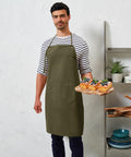 Man wearing a striped shirt and green apron holds a tray of assorted Premier PR181 sliced pastries with fruit toppings. Shelving with dishes and plants in the background.