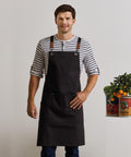 A man wears a Premier Clip n Clasp cross-back bib apron over a striped shirt indoors, standing with one hand on his hip beside tins, herbs, and a small potted orange tree arranged on a white surface.