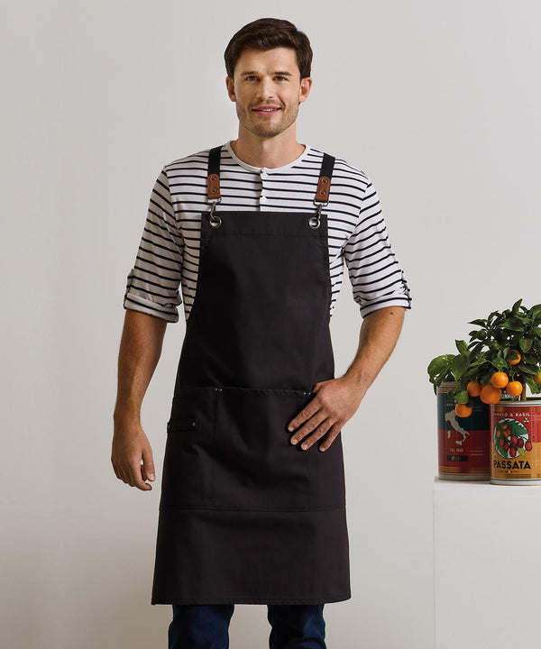 A man wears a Premier Clip n Clasp cross-back bib apron over a striped shirt indoors, standing with one hand on his hip beside tins, herbs, and a small potted orange tree arranged on a white surface.